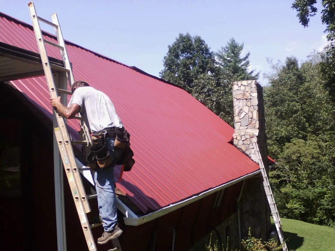 Roofer on a ladder beside a red metal roof and stone chimney — Soffit & Fascia Repair by Roofing Brothers in Fairfield